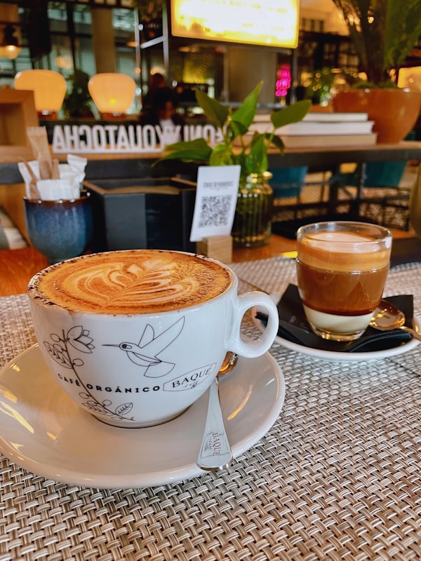 A beautifully presented cappuccino in a decorative cup sits on a woven placemat, accompanied by a glass of layered espresso on a black saucer. The background features a cozy caf&eacute; ambiance with warm lighting, potted plants, and a QR code on a menu stand.