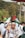 A crowd is gathered outdoors, with many people wearing headbands and scarves. The central figure has a 'SAVE GAZA' headband and a flag symbol on his cheek. Various flags are being held up in the background, and some people are taking photos. The environment is lush with greenery and palm trees.