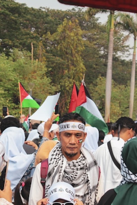 A crowd is gathered outdoors, with many people wearing headbands and scarves. The central figure has a 'SAVE GAZA' headband and a flag symbol on his cheek. Various flags are being held up in the background, and some people are taking photos. The environment is lush with greenery and palm trees.