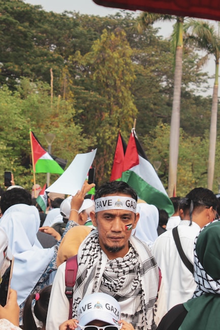 A crowd is gathered outdoors, with many people wearing headbands and scarves. The central figure has a 'SAVE GAZA' headband and a flag symbol on his cheek. Various flags are being held up in the background, and some people are taking photos. The environment is lush with greenery and palm trees.