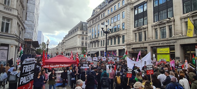A large crowd of people gathered on a city street, holding signs and banners advocating for the freedom of Palestine. The street is surrounded by tall buildings, including the University of Westminster. A red canopy labeled 'Socialist Party' is prominent among the crowd.