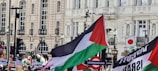 A crowd of people holding various flags and banners in front of a historical building with ornate architecture. Prominent among the flags is one with red, green, white, and black stripes, resembling the Palestinian flag. The banners contain slogans related to political causes, such as 'Free Palestine.' The setting appears to be an urban area with street lamps and a visible underground sign.
