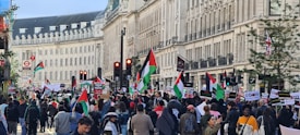 A large group of people are gathered on a city street, participating in a protest. They are holding signs and banners, and many are waving flags that are red, green, and black. The scene is set against a backdrop of historical buildings with distinct architectural features, and there are traffic lights visible around the area.
