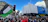 A large crowd gathers in a city street, holding signs and flags during a protest. The protestors appear passionate, with banners addressing political issues. The backdrop features urban architecture, a traffic light, and clear blue skies.