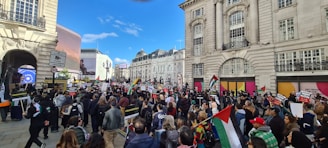 A large crowd of people gathers in an urban setting surrounded by historic buildings. Many individuals hold flags and protest signs with various messages. The scene is bustling with activity as the crowd moves through the city street, visible under a clear blue sky.