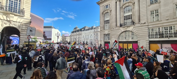 A large crowd of people gathers in an urban setting surrounded by historic buildings. Many individuals hold flags and protest signs with various messages. The scene is bustling with activity as the crowd moves through the city street, visible under a clear blue sky.