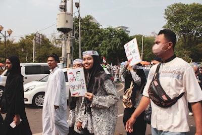 A group of people, some wearing traditional Muslim clothing and headbands reading 'Free Palestine,' participate in a protest or rally. They hold signs with messages supporting Palestine. The scene is outdoors, with trees and vehicles visible in the background.