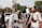 A group of people, some wearing traditional Muslim clothing and headbands reading 'Free Palestine,' participate in a protest or rally. They hold signs with messages supporting Palestine. The scene is outdoors, with trees and vehicles visible in the background.