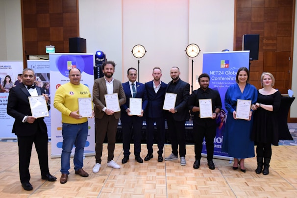 A group of eight people is standing indoors on a wooden floor, each holding a certificate. They are dressed in formal or semi-formal attire, including suits and dresses. Behind them, there are banners related to a conference and photography equipment like lights.