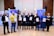 A group of eight people is standing indoors on a wooden floor, each holding a certificate. They are dressed in formal or semi-formal attire, including suits and dresses. Behind them, there are banners related to a conference and photography equipment like lights.