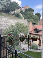 A charming courtyard scene featuring a stone pathway and several hanging flower pots with red blossoms. The area is enclosed by a black wrought iron fence. The background consists of a stone retaining wall covered with lush green ivy and other vegetation. Above, a couple of house roofs with red tiles are visible against a bright blue sky with scattered clouds. The overall scene conveys a peaceful and picturesque residential setting.