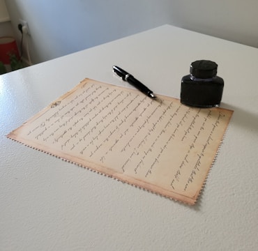 A piece of parchment paper with elegant handwritten text is placed on a table. Next to it, there's a black fountain pen and an ink bottle. The parchment is slightly tinted, giving it a vintage appearance. Light from the side casts subtle shadows, adding depth to the scene.