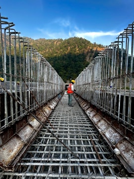A construction worker wearing a safety helmet and vest stands on a metal framework, which appears to be the unfinished structure of a bridge or large building. The scene is set outdoors with a backdrop of lush green hills under a clear blue sky. The metal bars are arranged in a grid pattern, indicating the early stages of the construction process.