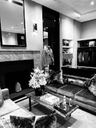 A sophisticated interior scene with a modern living room. The room features a sleek, marble fireplace, a large mirror above it, and a luxurious sofa adorned with plush velvet cushions. A coffee table is positioned centrally with a bouquet of flowers, a book titled Tom Ford 002, and two bottles on a tray. On the left side, there is a stylish jacket on a stand, with an open wardrobe showcasing neatly arranged clothing and bags.