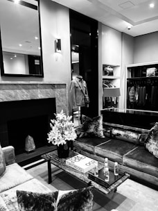A sophisticated interior scene with a modern living room. The room features a sleek, marble fireplace, a large mirror above it, and a luxurious sofa adorned with plush velvet cushions. A coffee table is positioned centrally with a bouquet of flowers, a book titled Tom Ford 002, and two bottles on a tray. On the left side, there is a stylish jacket on a stand, with an open wardrobe showcasing neatly arranged clothing and bags.