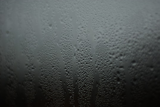 Water droplets streak down a glass surface, forming intricate patterns. The condensation creates a textured, misty appearance.