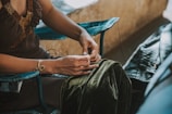 A person is seated, focusing on their hands while handling threads. They are wearing a brown dress adorned with a beaded neckline and a spiral bracelet. The person is sitting on a blue chair with a velvet fabric draped over their lap, and there is a leather item to the side.