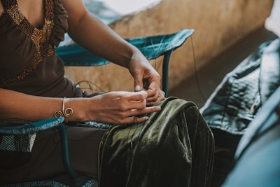 A person is seated, focusing on their hands while handling threads. They are wearing a brown dress adorned with a beaded neckline and a spiral bracelet. The person is sitting on a blue chair with a velvet fabric draped over their lap, and there is a leather item to the side.