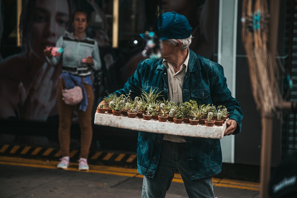 a man walking down a street carrying a tray of plants