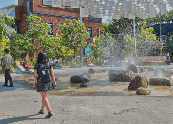 A public park area with water features where people are seen walking and relaxing. Large stones are set within shallow pools, creating a calming water display with mist rising in the air. Surrounding the water feature are trees and benches where some individuals are seated. A modern building with red brick and wood accents is visible in the background.