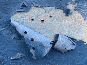 A close-up view of a wall with peeling blue paint, revealing a lighter surface beneath. The texture of the paint is rough, with curls and cracks creating an aged appearance. Multiple small holes are visible in the underlying layer.