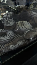 An assortment of artisanal breads is displayed inside a glass case. The breads are decorated with intricate patterns of flour and some have decorative scoring on their crusts. The scene includes various shapes such as round loaves, baguettes, and a wreath-shaped bread, all with a dark brown crust and a dusting of flour.