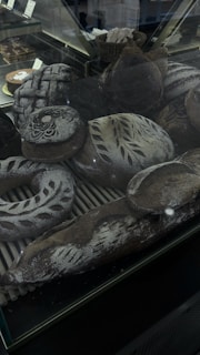 An assortment of artisanal breads is displayed inside a glass case. The breads are decorated with intricate patterns of flour and some have decorative scoring on their crusts. The scene includes various shapes such as round loaves, baguettes, and a wreath-shaped bread, all with a dark brown crust and a dusting of flour.