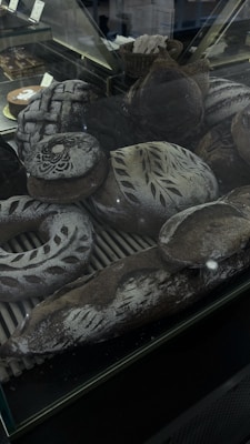 An assortment of artisanal breads is displayed inside a glass case. The breads are decorated with intricate patterns of flour and some have decorative scoring on their crusts. The scene includes various shapes such as round loaves, baguettes, and a wreath-shaped bread, all with a dark brown crust and a dusting of flour.