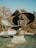 An abstract, metallic sculpture stands prominently on a stone pedestal in a landscaped garden. The sculpture features interconnected, swirling shapes and appears to be weathered. Surrounding the sculpture are autumn-colored trees and trimmed grass, with a stone balustrade in the background.
