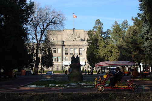A large government-style building with a flag atop is surrounded by trees. There is a statue in front of the building, and a person in a pedicab rides in the foreground. The scene is set on a clear day with blue skies.