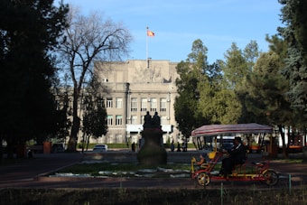 A large government-style building with a flag atop is surrounded by trees. There is a statue in front of the building, and a person in a pedicab rides in the foreground. The scene is set on a clear day with blue skies.