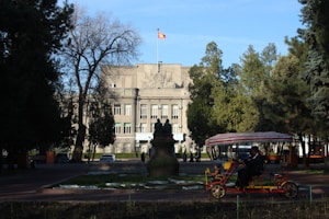 A large government-style building with a flag atop is surrounded by trees. There is a statue in front of the building, and a person in a pedicab rides in the foreground. The scene is set on a clear day with blue skies.