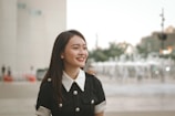 Young woman with long dark hair, dressed in a black outfit with a white collar and buttons, smiling while standing outdoors. A blurred background featuring buildings, people, and a fountain.