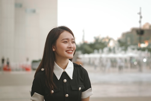 Young woman with long dark hair, dressed in a black outfit with a white collar and buttons, smiling while standing outdoors. A blurred background featuring buildings, people, and a fountain.