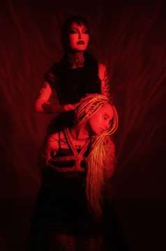 Two individuals are featured under a red-toned lighting. The person standing wears dark clothing and makeup, while the seated individual has long, light-colored braided hair and is wrapped in rope. The scene is dramatic with strong shadows.