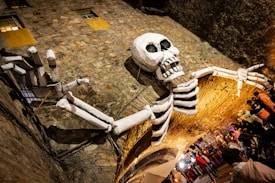 A large skeletal figure decorates an arched stone tunnel. The skeleton is attached to the wall, its long arms spread wide across the structure. Below, a crowd of people is gathered, some wearing bright colors, suggesting a lively event or festival.