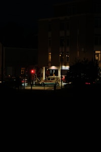 Nighttime image of a veterinary emergency clinic with a brightly lit entrance. There is a red traffic light in front of the building, and a white vehicle with 'VET' written on it is parked nearby. The scene appears calm with minimal activity.