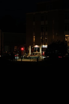 Nighttime image of a veterinary emergency clinic with a brightly lit entrance. There is a red traffic light in front of the building, and a white vehicle with 'VET' written on it is parked nearby. The scene appears calm with minimal activity.