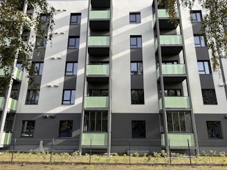 A modern residential building with several floors, featuring green-tinted glass balconies and a clean facade in shades of white and gray. The building has numerous windows, and there are trees partially visible around it, casting shadows on the structure. A fence surrounds the property, and there is a small patch of grass and plants at the base.