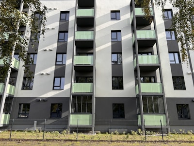 A modern residential building with several floors, featuring green-tinted glass balconies and a clean facade in shades of white and gray. The building has numerous windows, and there are trees partially visible around it, casting shadows on the structure. A fence surrounds the property, and there is a small patch of grass and plants at the base.