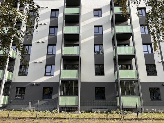 A modern residential building with several floors, featuring green-tinted glass balconies and a clean facade in shades of white and gray. The building has numerous windows, and there are trees partially visible around it, casting shadows on the structure. A fence surrounds the property, and there is a small patch of grass and plants at the base.
