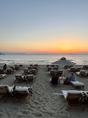 A serene beach scene with travelers relaxing on lounge chairs during sunset.