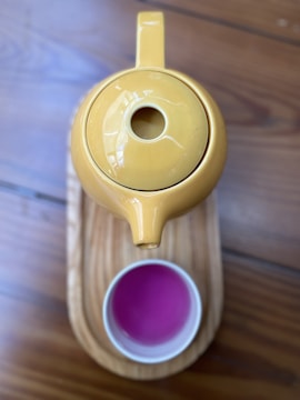 A yellow teapot is placed on top of a wooden tray, with a cup filled with a vibrant pink liquid below it. The setting is on a wooden surface with a warm tone.