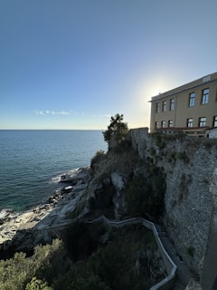 A large stone building is positioned on the edge of a steep cliff overlooking a calm sea. The sky is clear with sunlight creating a halo effect around the building. A winding stone path leads down the rocky cliffside, surrounded by patches of vegetation.