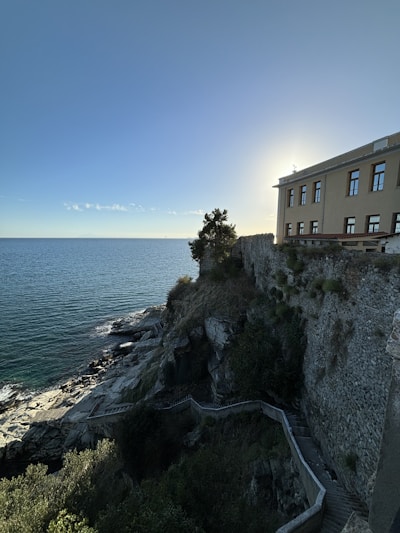 A large stone building is positioned on the edge of a steep cliff overlooking a calm sea. The sky is clear with sunlight creating a halo effect around the building. A winding stone path leads down the rocky cliffside, surrounded by patches of vegetation.
