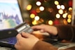 A person is using a laptop and holding a credit card, suggesting online shopping or making a payment. In the background, there is a display of blurred lights resembling a festive decoration.
