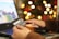 A person is using a laptop and holding a credit card, suggesting online shopping or making a payment. In the background, there is a display of blurred lights resembling a festive decoration.