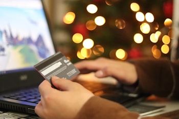 A person is using a laptop and holding a credit card, suggesting online shopping or making a payment. In the background, there is a display of blurred lights resembling a festive decoration.