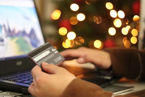 A person is using a laptop and holding a credit card, suggesting online shopping or making a payment. In the background, there is a display of blurred lights resembling a festive decoration.