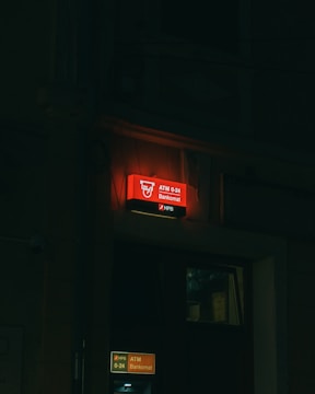 A dimly lit street scene featuring a glowing red ATM sign with the label 'Bankomat' and 'ATM 0-24'. The sign is positioned above the entrance to what appears to be a bank or financial institution, with another illuminated sign below. The overall setting is dark, suggesting nighttime.